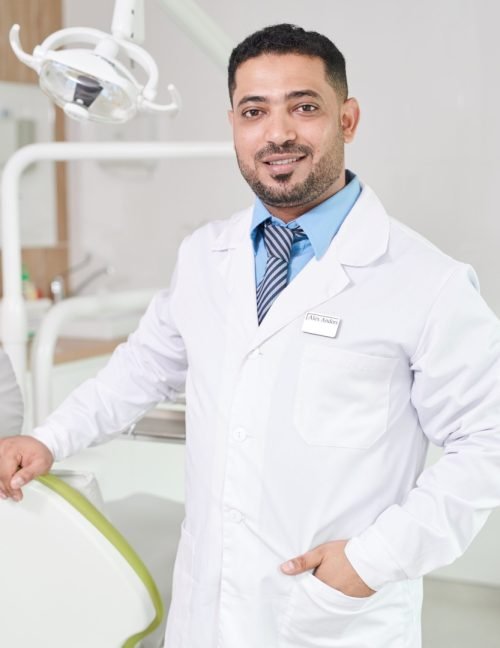 Portrait of Middle-Eastern dentist posing in office smiling at camera while standing by dental chair